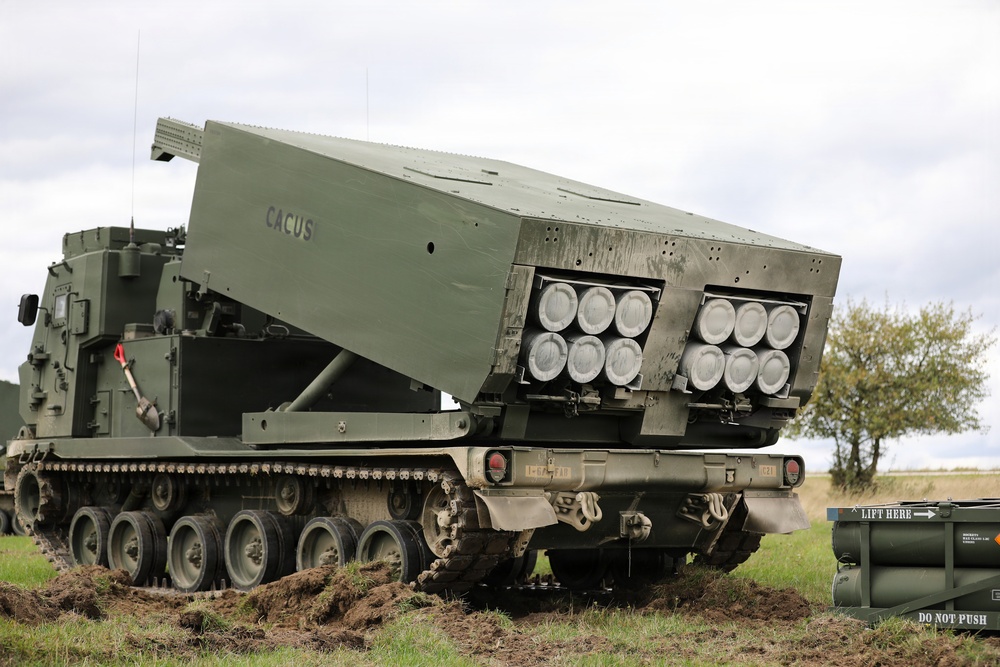 The image shows a tracked military multiple launch rocket system (MLRS) with its launcher pod elevated and angled, displaying two pods of rocket tubes (approximately 12 visible launch tubes total). The vehicle is marked with the name 'CACUS' on the launcher housing and is positioned on grass terrain with disturbed soil beneath the tracks. A reload container marked 'LIFT HERE' and 'DO NOT PUSH' is visible in the foreground right.