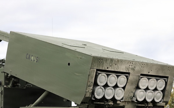 The image shows a tracked military multiple launch rocket system (MLRS) with its launcher pod elevated and angled, displaying two pods of rocket tubes (approximately 12 visible launch tubes total). The vehicle is marked with the name 'CACUS' on the launcher housing and is positioned on grass terrain with disturbed soil beneath the tracks. A reload container marked 'LIFT HERE' and 'DO NOT PUSH' is visible in the foreground right.