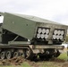 The image shows a tracked military multiple launch rocket system (MLRS) with its launcher pod elevated and angled, displaying two pods of rocket tubes (approximately 12 visible launch tubes total). The vehicle is marked with the name 'CACUS' on the launcher housing and is positioned on grass terrain with disturbed soil beneath the tracks. A reload container marked 'LIFT HERE' and 'DO NOT PUSH' is visible in the foreground right.