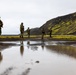 A group of approximately six armed soldiers in camouflage uniforms patrol across a dark volcanic terrain with vivid green moss-covered hillsides in the background. The landscape is characteristic of Iceland or a similar North Atlantic volcanic environment, featuring black soil, standing water puddles, and dramatic hillside contrasts. The soldiers' reflections are visible in a large foreground puddle, adding a compositional element to the image.