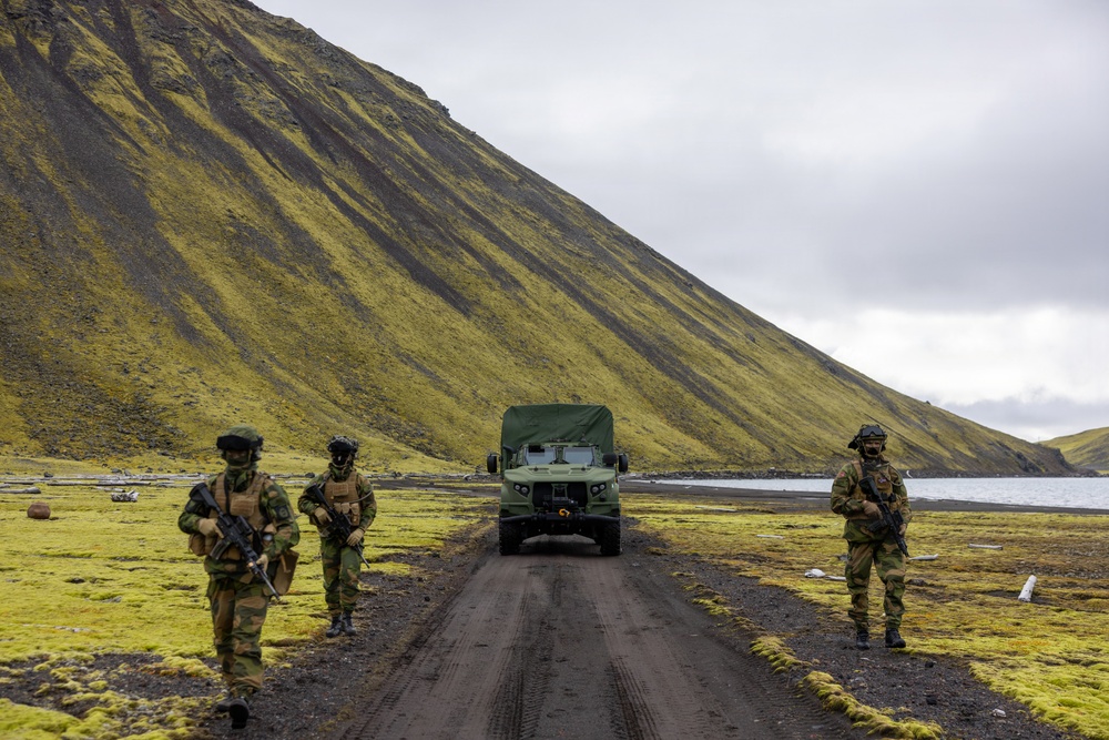 Three armed soldiers in camouflage uniforms are positioned along a dirt road in a rugged, moss-covered volcanic landscape, with a military truck stopped behind them. The terrain features a steep green moss-covered hillside, dark volcanic soil, and a body of water visible in the background. The scene appears to depict a military patrol or security operation in a Nordic or sub-Arctic environment.