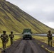 Three armed soldiers in camouflage uniforms are positioned along a dirt road in a rugged, moss-covered volcanic landscape, with a military truck stopped behind them. The terrain features a steep green moss-covered hillside, dark volcanic soil, and a body of water visible in the background. The scene appears to depict a military patrol or security operation in a Nordic or sub-Arctic environment.
