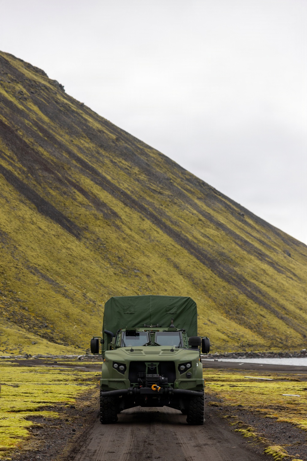 A military tactical vehicle, consistent with an Oshkosh JLTV (Joint Light Tactical Vehicle) in olive drab green, is parked or traveling on a dirt track in a remote, rugged landscape. The terrain features moss-covered volcanic hills and a small body of water in the background, strongly suggesting an Icelandic or North Atlantic environment. The vehicle is equipped with a front-mounted winch with a visible orange hook and a canvas cargo cover over the rear.