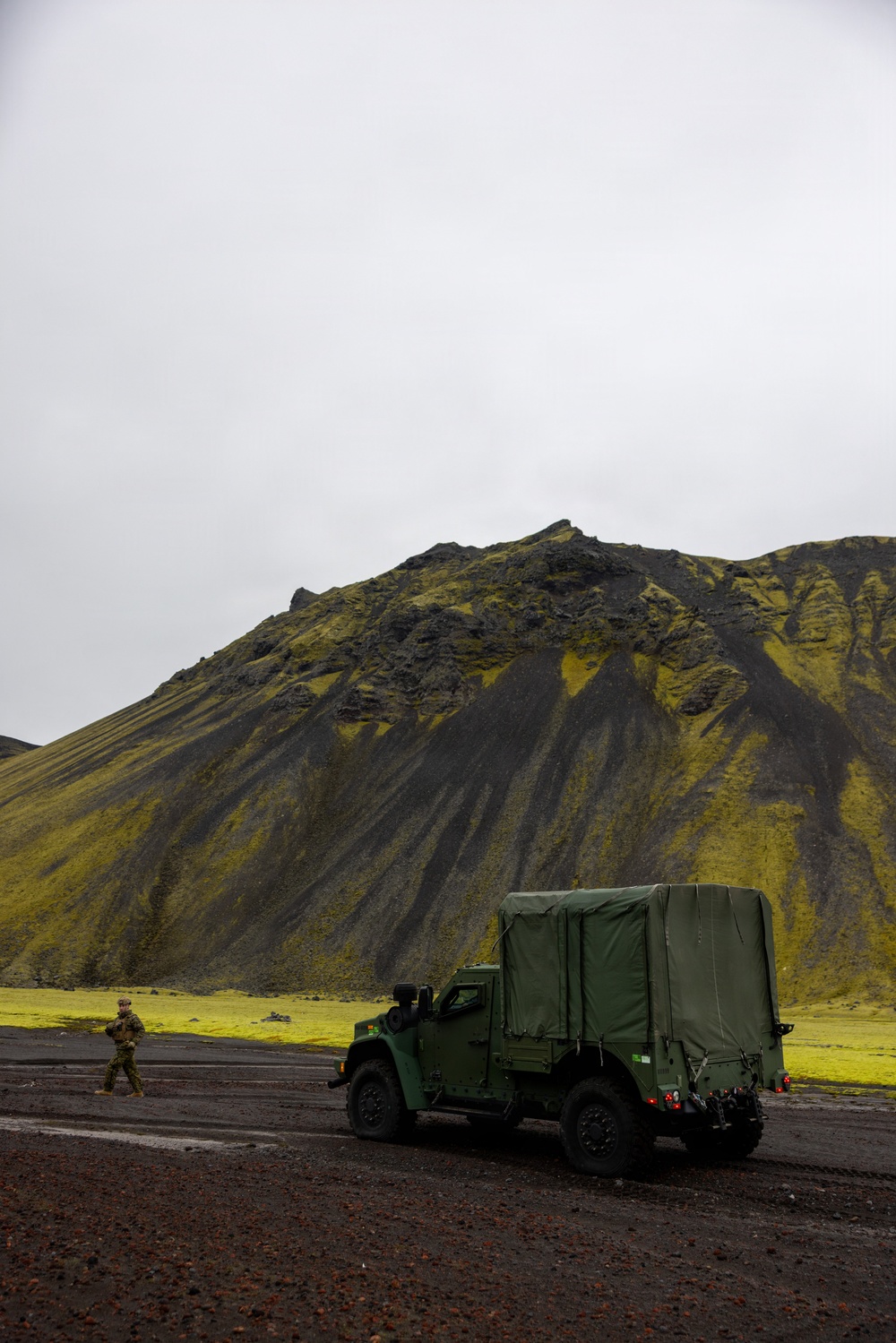 A military tactical vehicle with a canvas-covered cargo bed is parked on dark volcanic terrain in front of a dramatic moss-covered volcanic mountain. One soldier in camouflage uniform is visible walking near the vehicle. The landscape is consistent with Icelandic highland terrain, characterized by black volcanic rock and vivid green moss.