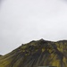 A military tactical vehicle with a canvas-covered cargo bed is parked on dark volcanic terrain in front of a dramatic moss-covered volcanic mountain. One soldier in camouflage uniform is visible walking near the vehicle. The landscape is consistent with Icelandic highland terrain, characterized by black volcanic rock and vivid green moss.