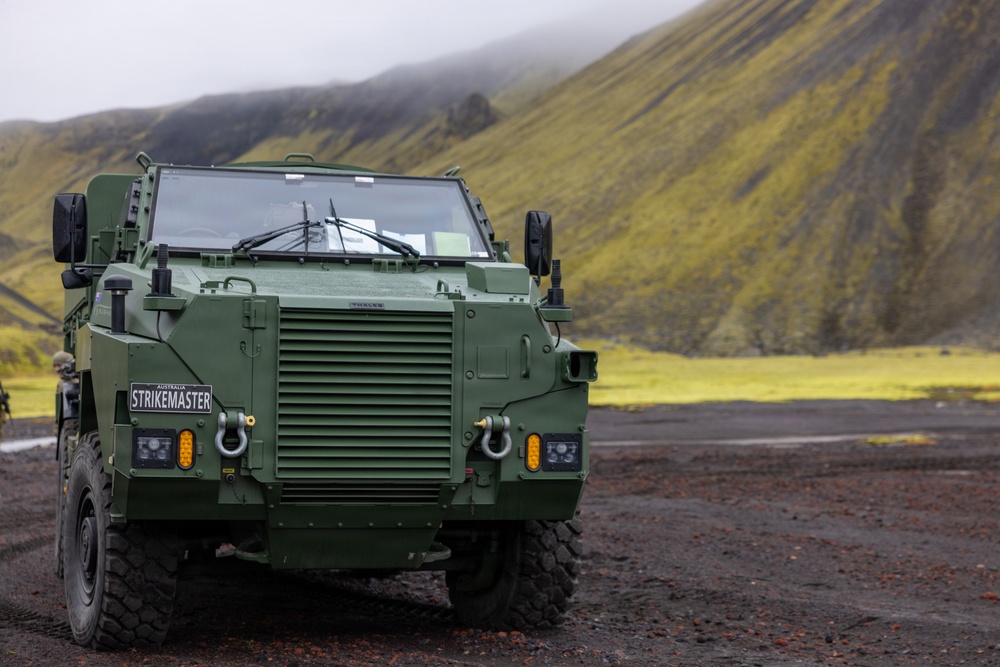 A military armored vehicle designated 'STRIKEMASTER' from Australia, manufactured by Thales, is parked on dark gravel terrain. The vehicle is painted in olive/military green and appears to be a protected mobility vehicle. The backdrop features dramatic green moss-covered volcanic hills partially obscured by low cloud or fog, consistent with an Icelandic or similar Nordic landscape.