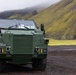 A military armored vehicle designated 'STRIKEMASTER' from Australia, manufactured by Thales, is parked on dark gravel terrain. The vehicle is painted in olive/military green and appears to be a protected mobility vehicle. The backdrop features dramatic green moss-covered volcanic hills partially obscured by low cloud or fog, consistent with an Icelandic or similar Nordic landscape.