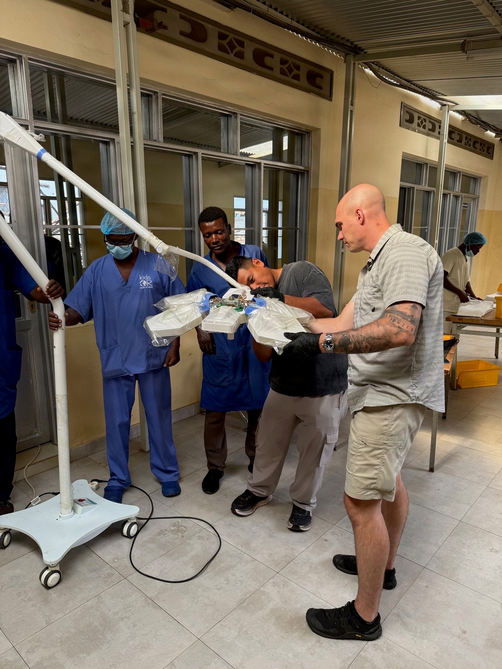 A group of individuals, including medical staff in blue scrubs and surgical caps/masks, are assembling or installing a mobile surgical/medical light fixture in what appears to be a hospital or clinic corridor. Two civilians in casual clothing are assisting with the setup, with one closely inspecting the light head assembly. The setting appears to be a medical facility in a developing country, possibly in Africa, based on the architecture and context.