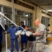 A group of individuals, including medical staff in blue scrubs and surgical caps/masks, are assembling or installing a mobile surgical/medical light fixture in what appears to be a hospital or clinic corridor. Two civilians in casual clothing are assisting with the setup, with one closely inspecting the light head assembly. The setting appears to be a medical facility in a developing country, possibly in Africa, based on the architecture and context.