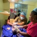 A medical team of four healthcare workers is attending to a newborn infant on a resuscitation warmer in what appears to be a clinical setting. The team includes at least one military medical personnel wearing camouflage uniform with sergeant rank insignia, alongside civilian medical staff. The infant is receiving immediate post-delivery care with tubing and medical interventions visible.
