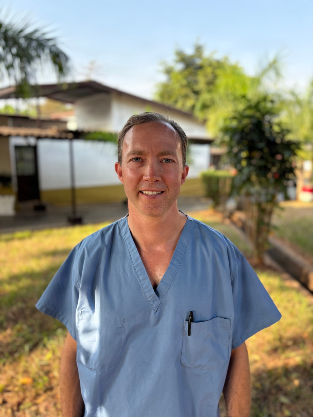 A man wearing light blue medical scrubs stands outdoors, smiling at the camera. He has a pen clipped to his chest pocket, suggesting a medical or healthcare professional role. The background shows a low-rise building, green trees, and a grassy area under a clear blue sky.