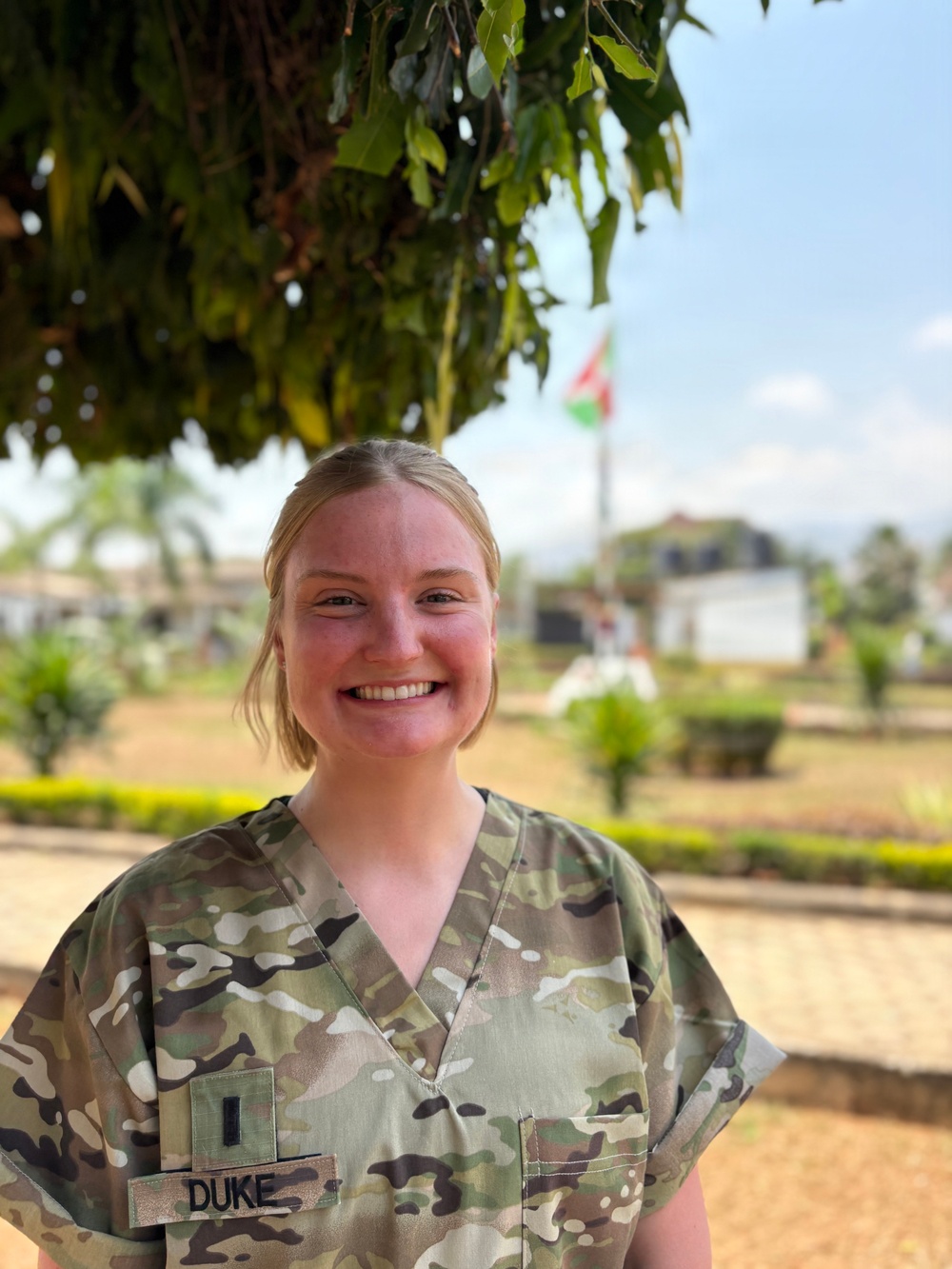 A young woman wearing military camouflage scrubs with a name tape reading 'DUKE' and a rank insignia patch (1st Lieutenant bar) is smiling at the camera. She is standing outdoors in what appears to be a tropical or subtropical environment, with a flag visible in the background that resembles the flag of Burundi. The setting suggests a military medical or humanitarian mission context.