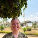 A young woman wearing military camouflage scrubs with a name tape reading 'DUKE' and a rank insignia patch (1st Lieutenant bar) is smiling at the camera. She is standing outdoors in what appears to be a tropical or subtropical environment, with a flag visible in the background that resembles the flag of Burundi. The setting suggests a military medical or humanitarian mission context.
