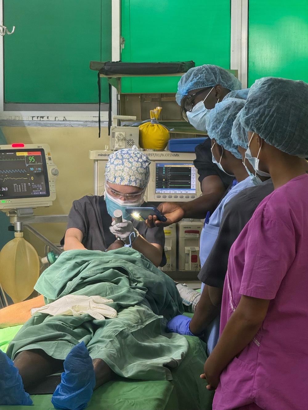 A medical procedure is being performed in what appears to be a surgical or clinical setting, likely in a resource-limited environment based on the green-painted walls and equipment visible. A central medical professional wearing a floral surgical cap, safety glasses, and mask is using a lighted laryngoscope or similar illuminated instrument on a draped patient. Two additional medical staff in scrubs and surgical caps observe the procedure, with patient monitoring equipment visible in the background.
