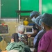 A medical procedure is being performed in what appears to be a surgical or clinical setting, likely in a resource-limited environment based on the green-painted walls and equipment visible. A central medical professional wearing a floral surgical cap, safety glasses, and mask is using a lighted laryngoscope or similar illuminated instrument on a draped patient. Two additional medical staff in scrubs and surgical caps observe the procedure, with patient monitoring equipment visible in the background.
