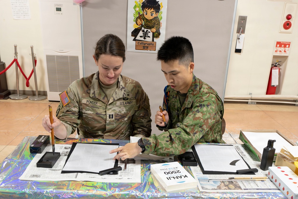 A U.S. Army soldier named Ponder, wearing captain rank insignia, participates in a Japanese calligraphy session alongside a Japan Ground Self-Defense Force soldier. They are seated at a table covered with calligraphy supplies, with a 'Calligraphy' sign visible in the background. This appears to be a cultural exchange activity between U.S. and Japanese military personnel.