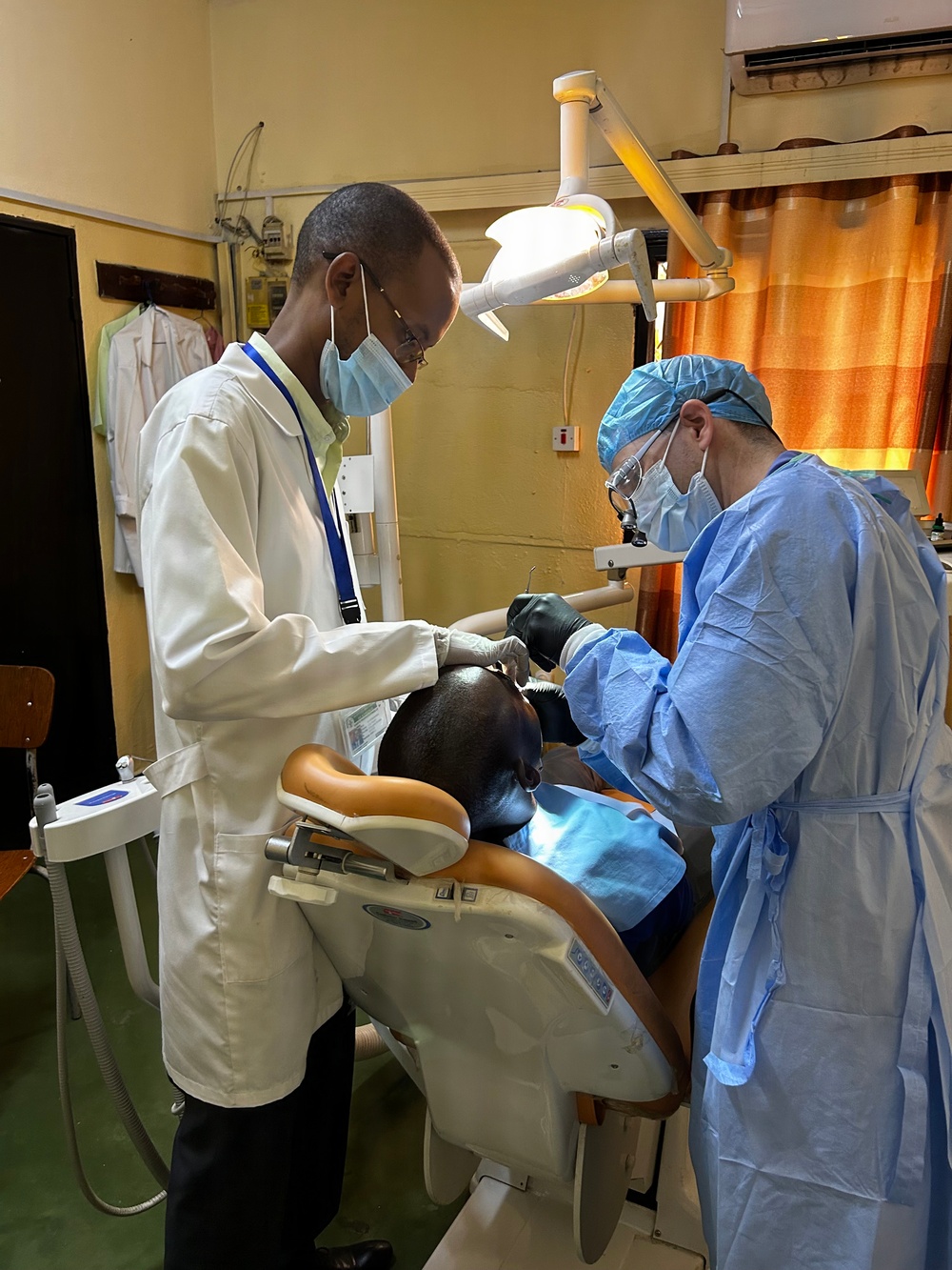 Two medical/dental professionals are performing a dental procedure on a seated patient in a clinical setting. One practitioner wears a white lab coat with a blue lanyard and surgical mask, while the other is fully gowned in blue surgical attire including cap, gown, mask, and black gloves. The setting appears to be a dental clinic in a resource-limited environment, suggested by the aged walls and basic furnishings.