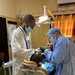 Two medical/dental professionals are performing a dental procedure on a seated patient in a clinical setting. One practitioner wears a white lab coat with a blue lanyard and surgical mask, while the other is fully gowned in blue surgical attire including cap, gown, mask, and black gloves. The setting appears to be a dental clinic in a resource-limited environment, suggested by the aged walls and basic furnishings.