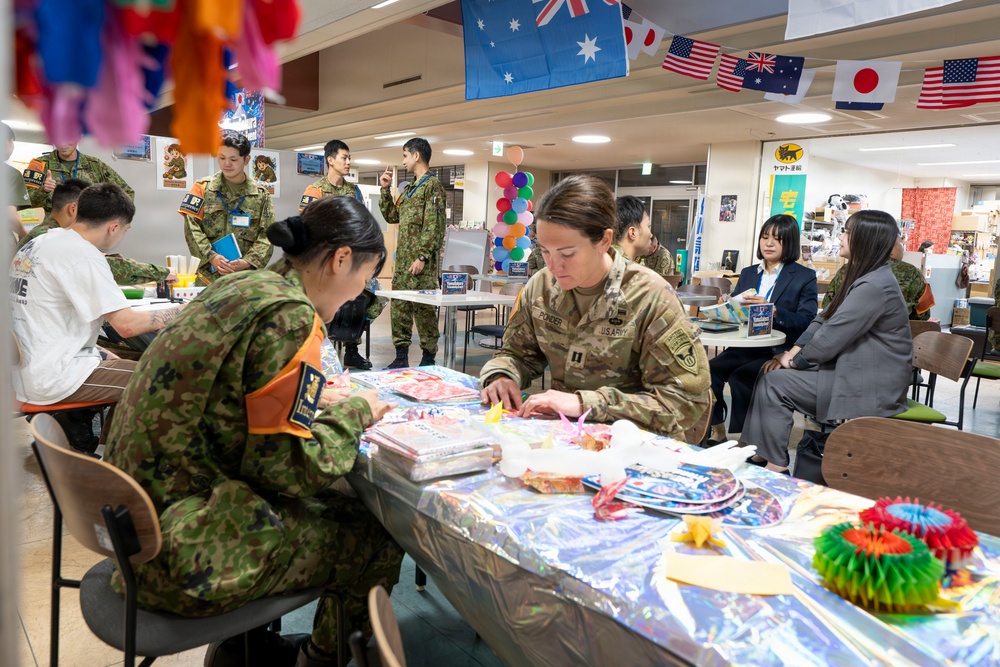 A multinational military gathering takes place in what appears to be a community or recreation room, with U.S. Army and Japanese Self-Defense Force personnel engaged in craft or activity tables. Flags from Australia, Japan, and the United States are visibly hanging from the ceiling, indicating an international cooperative event. Civilians in business attire are also present, suggesting a joint community engagement or cultural exchange activity.