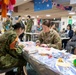 A multinational military gathering takes place in what appears to be a community or recreation room, with U.S. Army and Japanese Self-Defense Force personnel engaged in craft or activity tables. Flags from Australia, Japan, and the United States are visibly hanging from the ceiling, indicating an international cooperative event. Civilians in business attire are also present, suggesting a joint community engagement or cultural exchange activity.