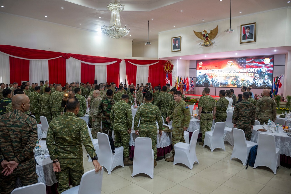 A military opening ceremony is taking place in a formal banquet hall decorated with red and white draping, consistent with Indonesian national colors. Multiple military personnel in various camouflage uniforms are gathered around round tables set with white chair covers, suggesting a multinational military exercise or conference. A large screen at the front displays 'THE OPENING CEREMONY' text along with what appears to be '2026' and military imagery.