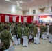 A military opening ceremony is taking place in a formal banquet hall decorated with red and white draping, consistent with Indonesian national colors. Multiple military personnel in various camouflage uniforms are gathered around round tables set with white chair covers, suggesting a multinational military exercise or conference. A large screen at the front displays 'THE OPENING CEREMONY' text along with what appears to be '2026' and military imagery.