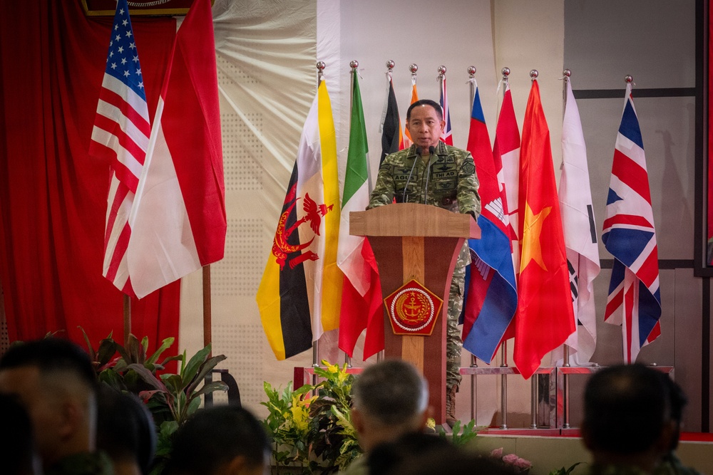 A military officer in camouflage uniform stands at a wooden podium bearing an emblem, delivering a speech at what appears to be a multinational military ceremony or conference. Multiple national flags are displayed behind him, suggesting an international military cooperation event. An audience of uniformed personnel is visible in the foreground.