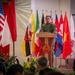 A military officer in camouflage uniform stands at a wooden podium bearing an emblem, delivering a speech at what appears to be a multinational military ceremony or conference. Multiple national flags are displayed behind him, suggesting an international military cooperation event. An audience of uniformed personnel is visible in the foreground.