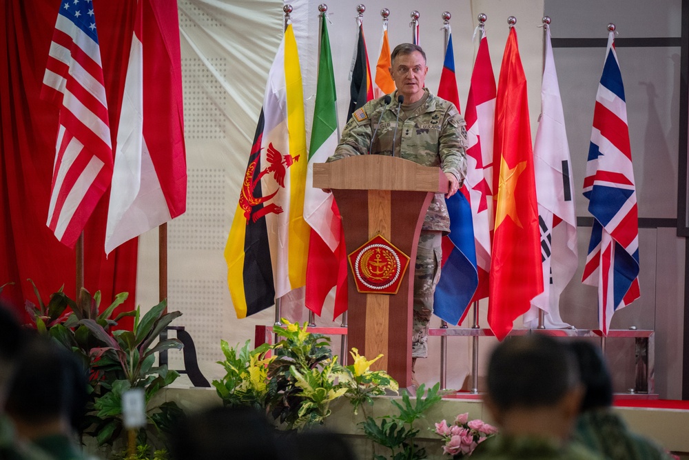 A military officer in camouflage uniform stands at a wooden podium delivering a speech at what appears to be a multinational military ceremony or conference. The backdrop features numerous national flags from various countries arranged in a row, with a prominent military emblem on the podium. An audience in military uniforms is partially visible in the foreground.