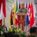 A military officer in camouflage uniform stands at a wooden podium delivering a speech at what appears to be a multinational military ceremony or conference. The backdrop features numerous national flags from various countries arranged in a row, with a prominent military emblem on the podium. An audience in military uniforms is partially visible in the foreground.