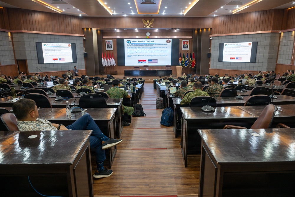 A military briefing or conference is taking place in a formal auditorium-style room, with attendees in various military uniforms and civilian attire seated at tiered desks. The central projection screen displays a presentation slide titled 'Road to Crisis (5 of 12)' featuring Indonesian and American flags, suggesting a joint military exercise or bilateral planning event. The setting appears to be an Indonesian military facility, evidenced by the Garuda Pancasila emblem and Indonesian flags visible on the front wall.