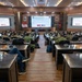 A military briefing or conference is taking place in a formal auditorium-style room, with attendees in various military uniforms and civilian attire seated at tiered desks. The central projection screen displays a presentation slide titled 'Road to Crisis (5 of 12)' featuring Indonesian and American flags, suggesting a joint military exercise or bilateral planning event. The setting appears to be an Indonesian military facility, evidenced by the Garuda Pancasila emblem and Indonesian flags visible on the front wall.
