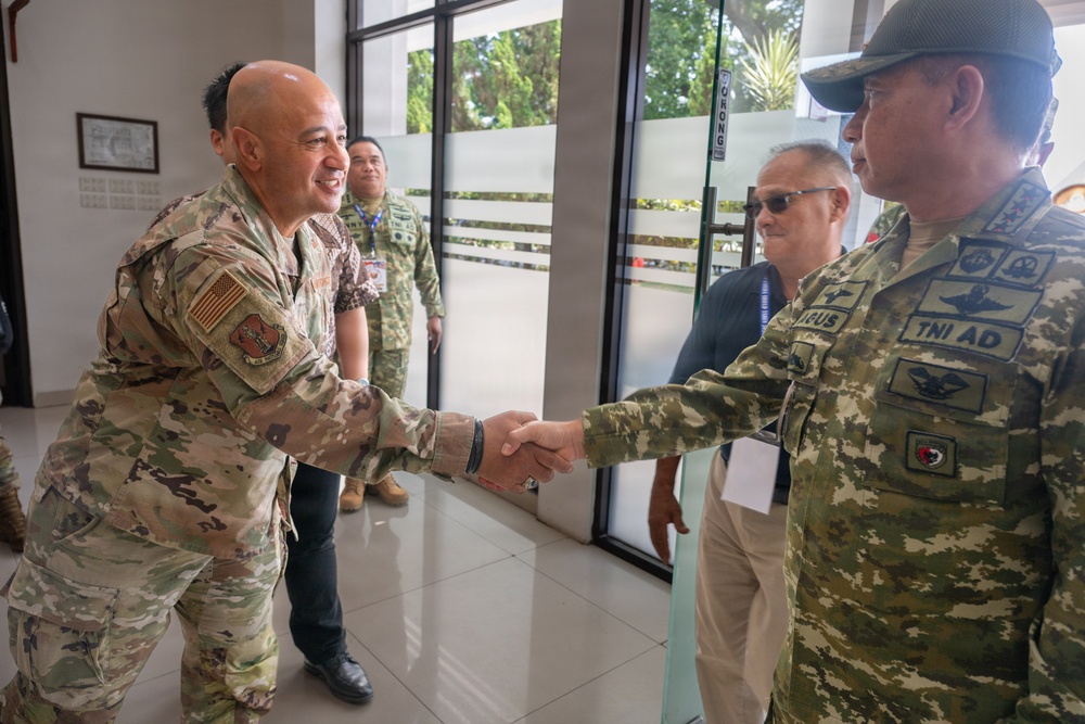 Two military personnel in camouflage uniforms are shaking hands in what appears to be an indoor facility, suggesting a formal military greeting or meeting. The individual on the left wears a US Army/Air Force OCP uniform with an American flag patch, while the individual on the right wears a uniform marked 'TNI AD', indicating Indonesian Army (Tentara Nasional Indonesia - Angkatan Darat). Several other individuals are visible in the background, suggesting a multilateral military engagement or liaison event.