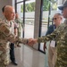 Two military personnel in camouflage uniforms are shaking hands in what appears to be an indoor facility, suggesting a formal military greeting or meeting. The individual on the left wears a US Army/Air Force OCP uniform with an American flag patch, while the individual on the right wears a uniform marked 'TNI AD', indicating Indonesian Army (Tentara Nasional Indonesia - Angkatan Darat). Several other individuals are visible in the background, suggesting a multilateral military engagement or liaison event.