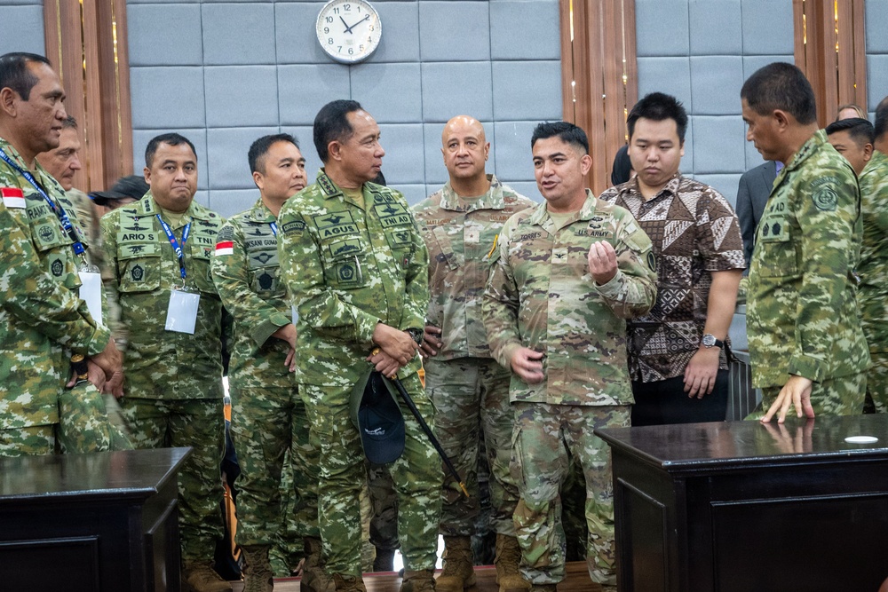 A group of military personnel from what appears to be the Indonesian Army (TNI AD) and the U.S. Army are gathered in a formal meeting room, engaged in discussion. Name tapes and patches visible on uniforms identify Indonesian soldiers (with 'TNI AD' markings) and at least two U.S. Army soldiers in MultiCam/OCP uniforms. The setting suggests a bilateral military engagement, briefing, or official visit.