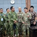 A group of military personnel from what appears to be the Indonesian Army (TNI AD) and the U.S. Army are gathered in a formal meeting room, engaged in discussion. Name tapes and patches visible on uniforms identify Indonesian soldiers (with 'TNI AD' markings) and at least two U.S. Army soldiers in MultiCam/OCP uniforms. The setting suggests a bilateral military engagement, briefing, or official visit.