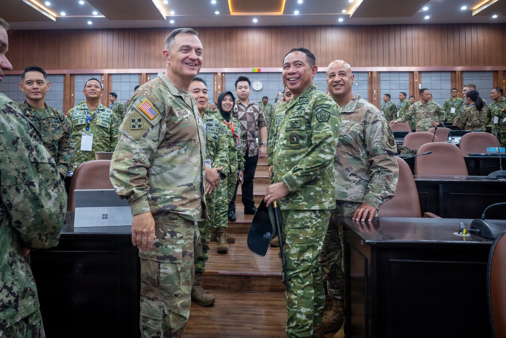 A group of military personnel from multiple nations are gathered in what appears to be a formal conference or briefing room. Two prominent figures in the foreground — one wearing a U.S. Army uniform with the name tape 'ELLIS' and an American flag patch, and another in a different camouflage pattern with 'TNI AD' (Indonesian Army) insignia — are smiling and interacting. The setting suggests a multinational military meeting or exercise.