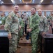 A group of military personnel from multiple nations are gathered in what appears to be a formal conference or briefing room. Two prominent figures in the foreground — one wearing a U.S. Army uniform with the name tape 'ELLIS' and an American flag patch, and another in a different camouflage pattern with 'TNI AD' (Indonesian Army) insignia — are smiling and interacting. The setting suggests a multinational military meeting or exercise.
