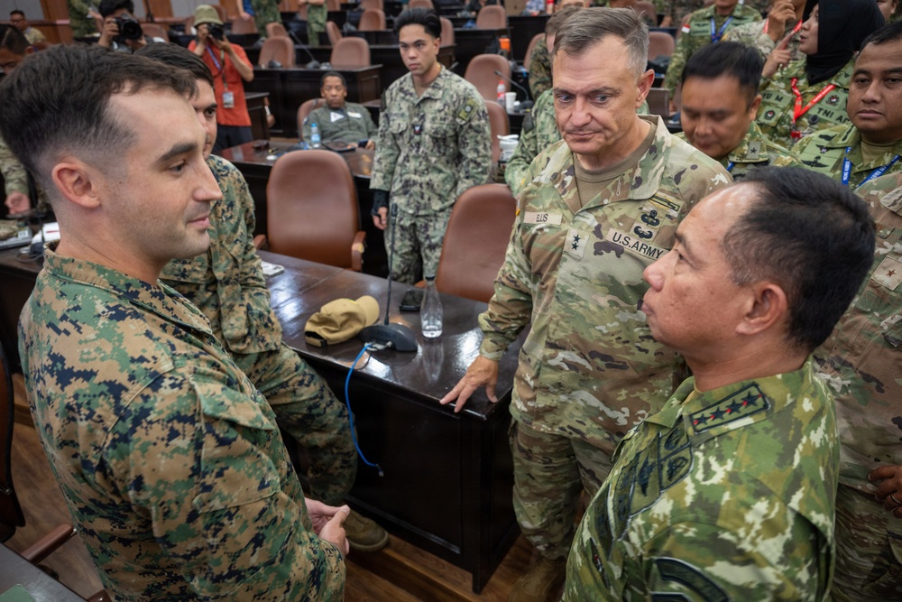 A multinational military gathering appears to be taking place in a large conference or assembly room. A U.S. Army officer with two stars (Major General) and the name tape 'ELIS' is engaged in conversation with personnel wearing different camouflage uniforms, including U.S. Marine Corps MARPAT and what appear to be Southeast Asian military uniforms. The setting suggests a joint military exercise, briefing, or multinational coordination event.