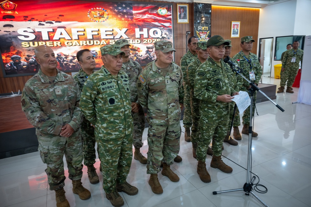 A group of military officers from multiple nations are gathered at the STAFFEX HQ for Super Garuda Shield 2025, a joint military exercise. Personnel wearing both US Army OCP uniforms and Indonesian TNI camouflage uniforms are standing together, with one officer appearing to deliver remarks at a microphone stand. The backdrop features the official exercise banner with both US and Indonesian flags.