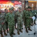 A group of military officers from multiple nations are gathered at the STAFFEX HQ for Super Garuda Shield 2025, a joint military exercise. Personnel wearing both US Army OCP uniforms and Indonesian TNI camouflage uniforms are standing together, with one officer appearing to deliver remarks at a microphone stand. The backdrop features the official exercise banner with both US and Indonesian flags.