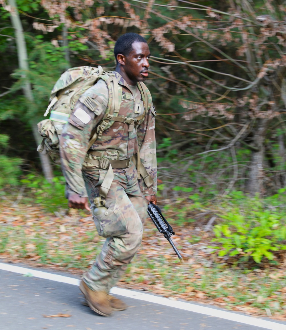 A U.S. Army soldier in OCP (Operational Camouflage Pattern) uniform is conducting a ruck march along a paved road bordered by woodland. The soldier is carrying a fully loaded rucksack and holding what appears to be an M4/M16-style rifle at his side. The image captures motion blur on the feet, indicating a brisk pace.