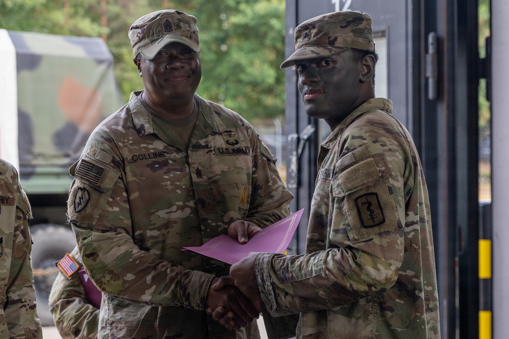 Two U.S. Army soldiers are shaking hands in what appears to be a formal recognition or award ceremony. The soldier on the left, identified by his name tape as 'Collins,' holds a pink/purple folder and appears to be presenting it to the other soldier. Both are wearing OCP (Operational Camouflage Pattern) uniforms with unit patches visible.