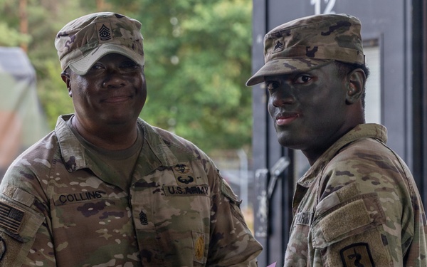 Two U.S. Army soldiers are shaking hands in what appears to be a formal recognition or award ceremony. The soldier on the left, identified by his name tape as 'Collins,' holds a pink/purple folder and appears to be presenting it to the other soldier. Both are wearing OCP (Operational Camouflage Pattern) uniforms with unit patches visible.