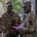Two U.S. Army soldiers are shaking hands in what appears to be a formal recognition or award ceremony. The soldier on the left, identified by his name tape as 'Collins,' holds a pink/purple folder and appears to be presenting it to the other soldier. Both are wearing OCP (Operational Camouflage Pattern) uniforms with unit patches visible.