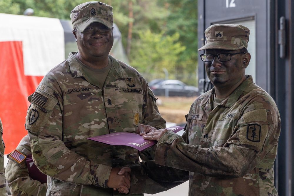 Two U.S. Army soldiers in OCP (Operational Camouflage Pattern) uniforms are shaking hands while jointly holding a purple/maroon folder or certificate holder, suggesting an award or recognition ceremony. The soldier on the left, identified by his name tape as 'COLLINS,' wears a Command Sergeant Major rank insignia, while the soldier on the right wears a Sergeant rank insignia and glasses. The setting appears to be an outdoor military installation, likely in Europe based on contextual background elements.