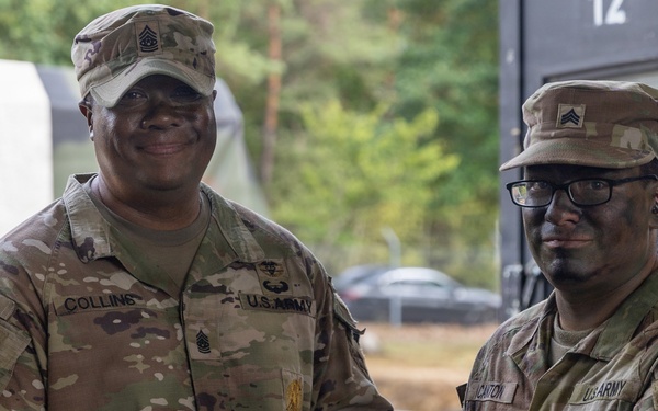 Two U.S. Army soldiers in OCP (Operational Camouflage Pattern) uniforms are shaking hands while jointly holding a purple/maroon folder or certificate holder, suggesting an award or recognition ceremony. The soldier on the left, identified by his name tape as 'COLLINS,' wears a Command Sergeant Major rank insignia, while the soldier on the right wears a Sergeant rank insignia and glasses. The setting appears to be an outdoor military installation, likely in Europe based on contextual background elements.