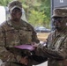 Two U.S. Army soldiers in OCP (Operational Camouflage Pattern) uniforms are shaking hands while jointly holding a purple/maroon folder or certificate holder, suggesting an award or recognition ceremony. The soldier on the left, identified by his name tape as 'COLLINS,' wears a Command Sergeant Major rank insignia, while the soldier on the right wears a Sergeant rank insignia and glasses. The setting appears to be an outdoor military installation, likely in Europe based on contextual background elements.