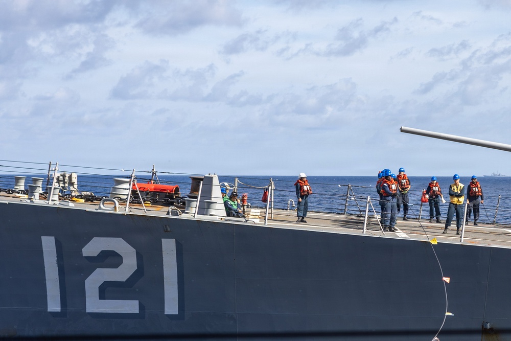 A naval vessel bearing hull number '121' is shown at sea with crew members stationed along the deck. Personnel are wearing orange life vests and blue hard hats, suggesting an underway replenishment or mooring operation is in progress. The ship appears to be a U.S. Navy guided-missile cruiser or destroyer operating in open ocean conditions.
