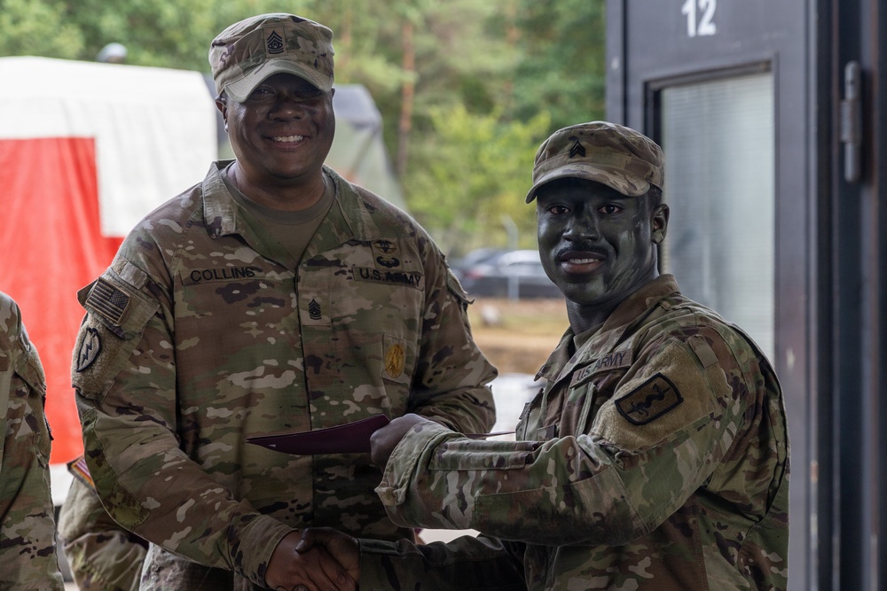 Two U.S. Army soldiers in OCP (Operational Camouflage Pattern) uniforms are posing together while shaking hands and exchanging what appears to be a certificate or document. The soldier on the left, identified by his name tape as 'Collins,' wears a Command Sergeant Major rank insignia on his cap, suggesting this is an award or recognition ceremony. The soldier on the right has tactical face paint applied, indicating recent or ongoing field training exercises.